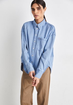 Camicia di jeans azzurro chiaro con bottoni, colletto, taschino sul petto e orlo curvo, abbinata a pantaloni color sabbia e anelli argentati sulle mani.
