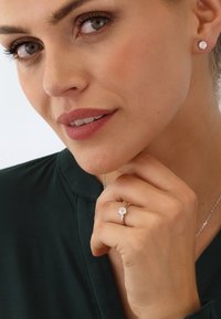 Silberring mit einem runden, klaren Edelstein, getragen an einer Hand mit lackierten Nägeln; kombiniert mit passenden Ohrsteckern und einer filigranen Silberkette.