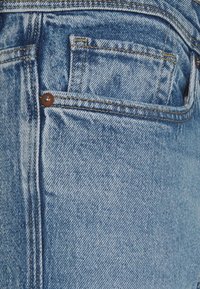 Blå denimjeans har en vanlig ficka med en brun knapp och sömnadsdetaljer. Tyget visar en klassisk blekt textur.