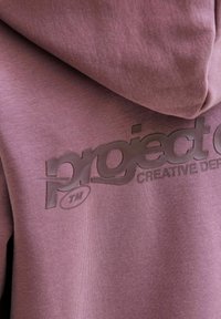 Togica s kapuljačom u umirujućoj ljubičastoj boji, sa teksturiranim materijalom i sjajnim crnim logotipom s natpisima "project" i "CREATIVE DE" na prednjoj strani.