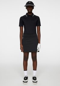 Svart pikétröja med vita detaljer, ihop med en svart kjol. Tillbehör inkluderar en svart visir och en vit golfhandske, bärs med svart och vita sneakers.