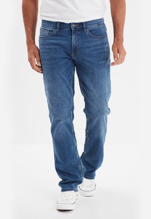Man draagt middenblauwe rechte jeans, witte sneakers en een wit overhemd, staand tegen een effen witte achtergrond.