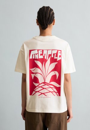 Personne aux cheveux tressés porte un T-shirt blanc avec un grand motif rouge d'ananas et le texte stylisé "Pineapple" au dos.