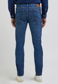 Jeans de ganga azul escura com um corte slim, apresentando um leve desgaste, dois bolsos traseiros e um patch de marca em pele castanha na cintura.