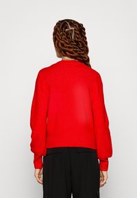 Henkilö, jolla on pitkät kiharaiset letit, pukeutuneena kirkkaan punaiseen neuleeseen ja mustiin housuihin, näkyy selkäpuolelta yksinkertaista taustaa vasten.