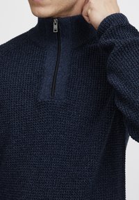 Pulover tricotat de culoare bleumarin, cu guler înalt și o fermoar scurt. Textura este groasă și țesută, având o suprafață texturată și mâneci lungi.