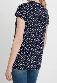 Marinblus med korta ärmar, prydd med ett vitt blommönster, har detaljer med rynkad yoke och en avslappnad passform, i kombination med blå jeans.