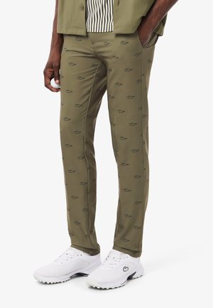Pantalones verde oliva con un patrón de logotipo en verde oscuro repetitivo, diseño ajustado y textura suave; combinados con zapatillas deportivas blancas.
