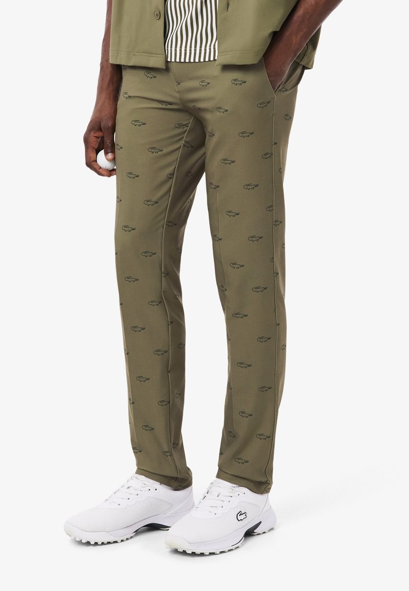 Pantalon vert olive avec un motif de logo vert foncé répétitif, coupe slim et texture lisse ; associé à des chaussures de sport blanches.
