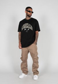 Čierne bavlnené tričko s nápisom "MIGONZALES" potlačeným v bielej a béžovej farbe, spárované s béžovými cargo nohavicami. Biely tenisky a čierne slnečné okuliare dotvárajú outfit.