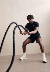 Maglietta e pantaloni corti neri da performance, con funi da battaglia nere spesse; scarpe da atletica bianche e gialle, ambientazione indoor con sfondo neutro chiaro.