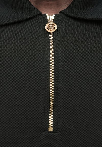 Juodas tekstūruotas polo marškinėliai su auksiniu užtrauktuku, kurio trauktukas yra liūto galvos formos. Apykaklė plokščia, nurodanti sportišką dizainą.