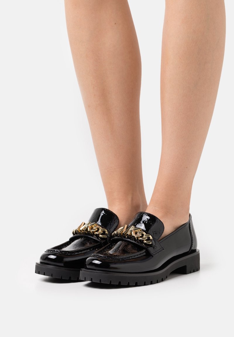Mocassins en cuir verni noir avec une semelle épaisse noire et un accent en chaîne dorée sur le cou-de-pied. Texture lisse avec une finition brillante.
