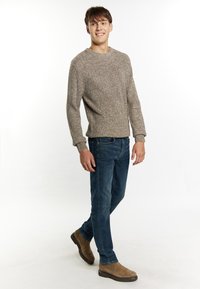 Pulover tricotat de culoare maro deschis, cu un model texturat, blugi blue jeans și cizme până la gleznă de culoare bej, stând pe un fundal alb simplu.