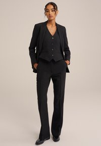 Černý oblek na míru obsahující přiléhavý blejzr, vestu s knoflíky a kalhoty rovného střihu. Hladká tkanina s elegantním povrchem.