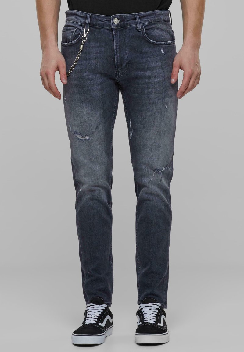 Tummansiniset denim-farkut slim fit -leikkauksella, kulutetut yksityiskohdat, ketjupidin ja napitus, yhdistettynä mustiin lenkkitossuihin.