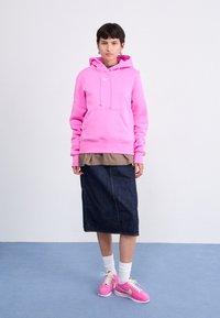 Roze hooded sweatshirt met een voorzak, gelaagd over een bruine top en een donkere spijkerrok. Gecombineerd met witte sokken en roze sneakers.