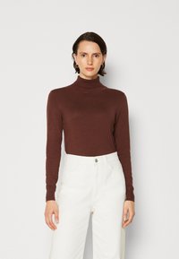 Hnědý dlouhý rukáv turtleneck svetr vyrobený z měkké tkaniny, s přiléhavými rukávy a žebrovaným límcem, v kombinaci s bílými kalhotami s vysokýmPasem.
