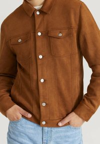 Chaqueta de ante marrón con un cuello clásico, botones de presión plateados, dos bolsillos en el pecho y una textura suave. Llevada sobre denim claro.