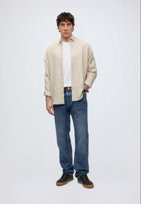 Jonge man staand, draagt een beige overhemd met knopen over een wit T-shirt, blauwe spijkerbroek, bruine riem, zwarte sneakers en een polshorloge.