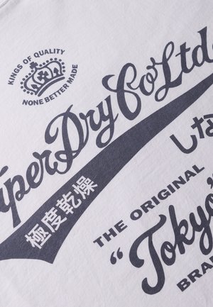 Camiseta de algodón blanca con un diseño gráfico en texto azul oscuro, que incluye "SuperDry Co Ltd" y un logo de corona, con caracteres japoneses incluidos.