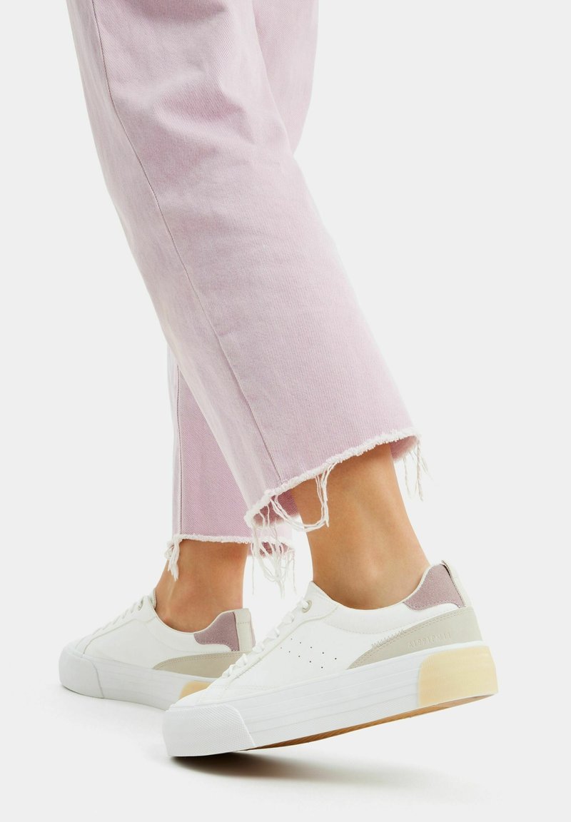 Białe sneakersy z pastelowymi akcentami, z teksturowaną skórzaną cholewką, perforacjami, masywną podeszwą i lekko postrzępionymi jasnoróżowymi dżinsami.