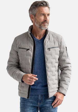 Mann trägt hellgraue gesteppte Jacke über blauem Pullover und Jeans, schaut zur Seite und stellt mit einer Hand den Reißverschluss der Jacke ein.