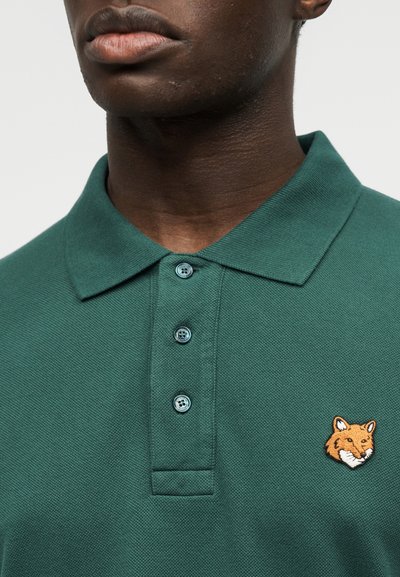 Gros plan sur une personne portant un polo vert foncé avec trois boutons et une tête de renard orange brodée sur le côté gauche de la poitrine.