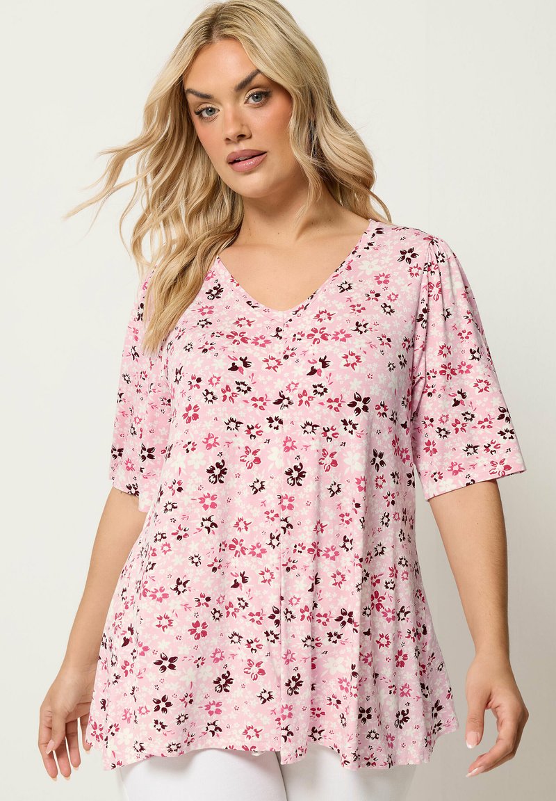 Femme blonde portant une tunique rose à fleurs avec un col en V et des manches aux coudes, debout devant un fond neutre uni.
