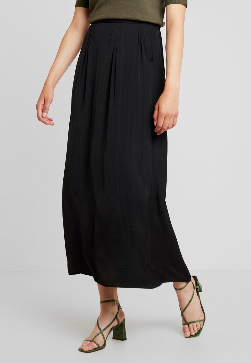 Femme portant une longue jupe noire plissée associée à des sandales vertes à talons blocs avec des brides et un haut vert olive, debout sur un sol gris.