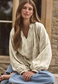 Hellbeige Bluse aus strukturiertem Stoff, mit fallenden Schultern und elastischen Bündchen. Kombiniert mit hoch taillierten Jeans im lässigen Schnitt.
