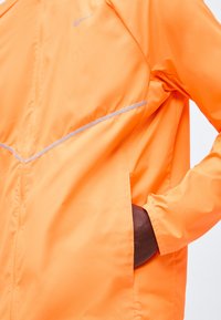 Bright orange Nike jacket with reflective stripe and logo, featuring a hand partially inside a side pocket.