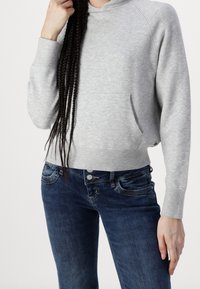 Personne portant un pull à manches longues gris clair et un jean bleu foncé, tenant une longue mèche de cheveux tressés d'une main.