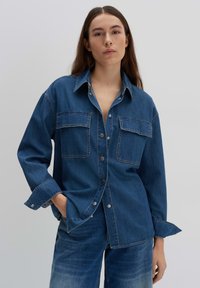 Camicia in jeans con bottone, dotata di due tasche sul petto, colletto e maniche lunghe. Colore: blu medio con cuciture a contrasto e bottoni metallici.
