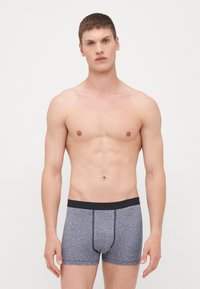 Grau Baumwoll-Boxershorts mit schwarzem Bund, mit einem figurbetonten Design und kontrastierenden Nähten. Das Modell steht mit den Armen an den Seiten.