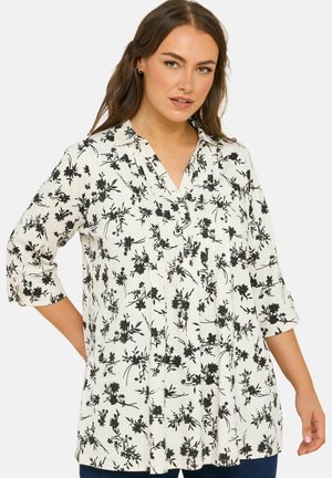 Vrouw draagt een witte blouse met zwarte bloemenprint en driekwart mouwen, poseert met één hand op haar heup tegen een effen achtergrond.