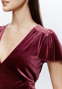Robe à col en V en velours bordeaux profond, dotée de manches bouffantes et d'un corsage ajusté avec un froncé subtil à la taille pour une silhouette structurée.