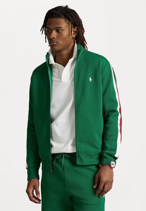 Veste zippée verte en coton, avec une accentuation à rayures blanches et rouges sur les manches, associée à un polo blanc et un pantalon vert assorti.