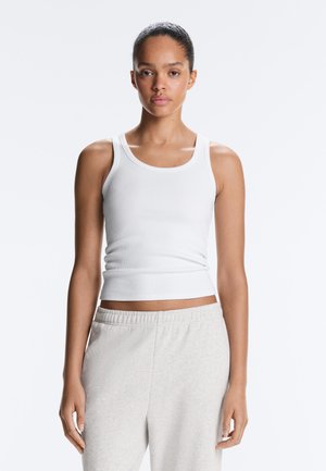 Femeie cu părul prins la spate, purtând un top alb fără mâneci cu dungi și pantaloni de trening gri deschis, stând în fața unui fundal simplu.