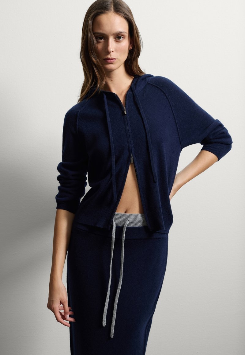 Dunkelblauer Reißverschluss-Hoodie aus weichem Material mit lockerer Passform. Verfügt über einen grauen Bund, Kordeln und ein verkürztes Design, kombiniert mit einem passenden Rock.