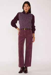 Gilet viola lavorato a maglia sopra una blusa bordeaux; abbinato a pantaloni in corda a gamba larga color prugna. Stivali alla caviglia marroni e accessori dorati.