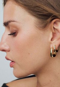 Guldtonade ringörhängen med en slät, glänsande yta, formade i en vågig design, bärs på en models öra mot en neutral bakgrund.