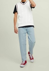 Weißer gestrickter ärmelloser Pullover mit Zopfmuster, getragen über einem schwarzen T-Shirt, kombiniert mit hellblauen Jeans und roten Turnschuhen.