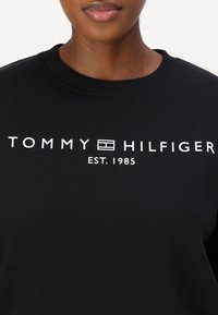 Sudadera negra con cuello redondo, que presenta texto blanco: "TOMMY HILFIGER EST. 1985". Diseño sencillo con una textura suave.
