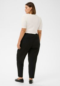 Femeie cu păr brunet până la umeri, purtând un tricou alb cu mânecă scurtă, pantaloni negri conici și loafers negri, stând pe un fundal alb.