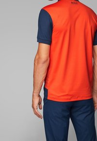 Červené a námořnické modré polo tričko s krátkým rukávem a texturovaným zadním dílem, zdobené úzkými pruhy a logem u výstřihu. Jednoduchý, sportovní design.