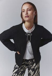 Black cardigan with a textured knit, featuring a bow detail at the neck. Worn over a white T-shirt. Paired with patterned pants.