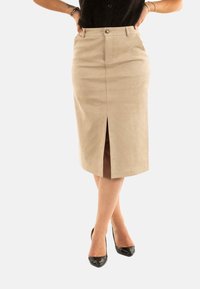 Femme portant une jupe crayon beige à mi-cuisse avec une fente avant, un haut sans manches noir et des escarpins noirs, se tenant avec les jambes croisées.