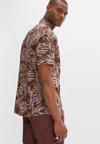 Cammicia a maniche corte marrone con motivo palme color crema, colletto a bottoni e texture liscia. Il modello indossa pantaloni marroni abbinati.