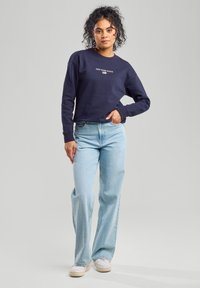 Marine sweatshirt met de tekst "NEW YORK STATE", gecombineerd met lichtblauwe wijde jeans en witte sneakers. Krullend haar, handen op de heupen.